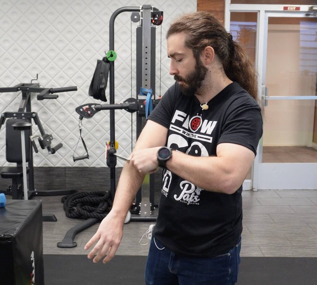 Dr. John standing holding a thin metal muscle scraping tool in his left hand applying pressure and scraping his right forearm just below the elbow